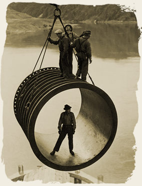 Four men, three standing on top and one standing inside, riding on large casing section of pipe suspended by cable as it is moved into position by a crane, reservoir in the background during construction of the Grand Coulee Dam, Washington, 1936-46